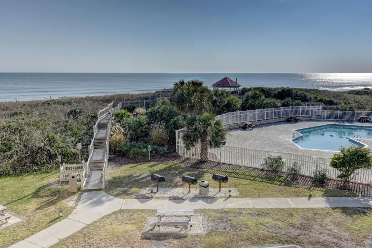 A sparkling swimming pool sits at the center, bordered by barbecue grills, picnic tables, and lush greenery. In the background, the ocean is visible, creating a seamless blend of poolside and beachfront leisure. The inviting setting is perfect for both relaxation and social gatherings, with multiple amenities on display.