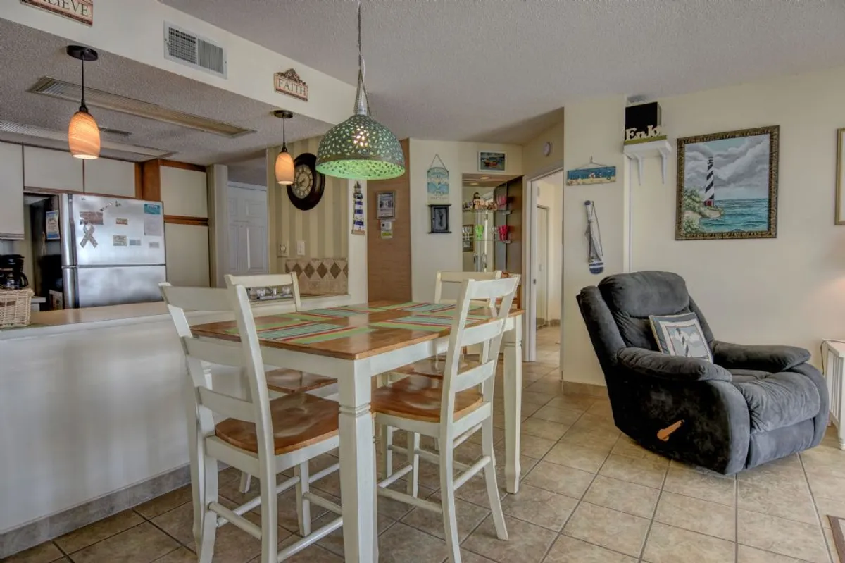 An open-plan room featuring a dining table next to a visible kitchen area, with comfortable seating and decorative accents. The space exudes comfort and homeliness, ideal for both meals and casual interactions. The kitchen is visible, tying the culinary and dining experience together.