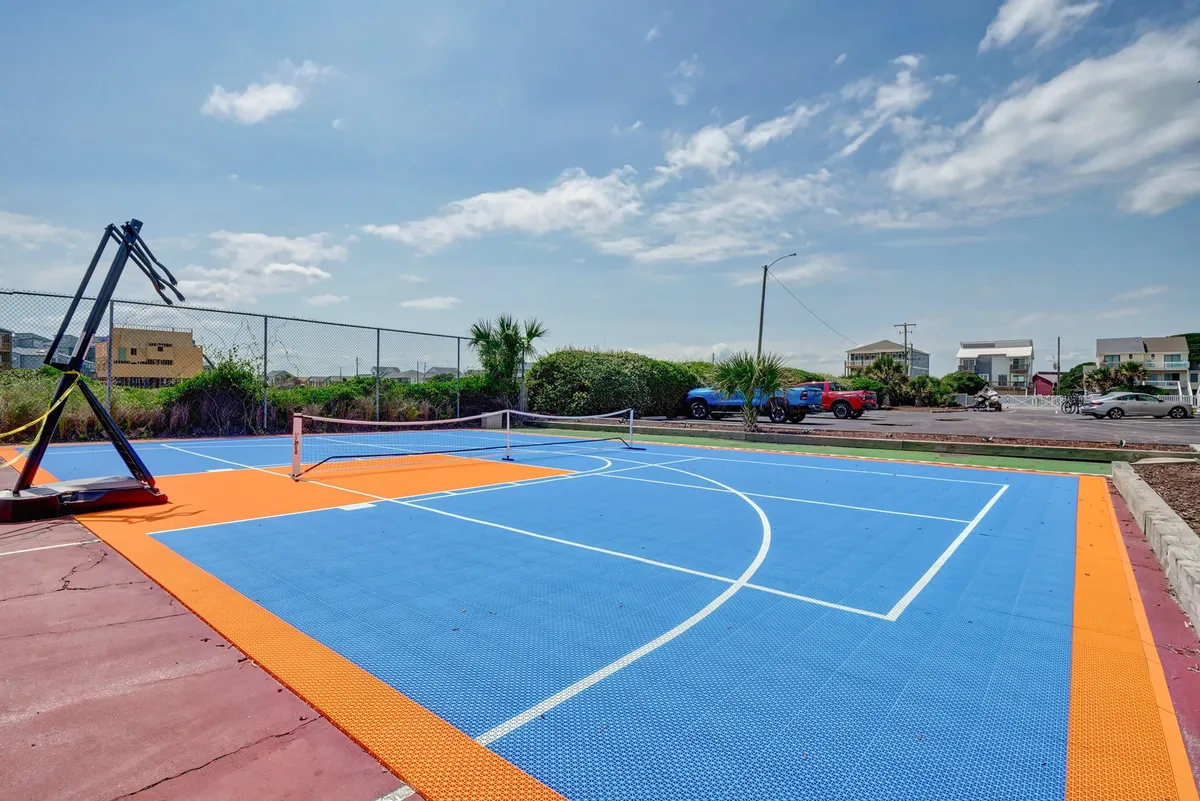 An exterior shot of a basketball court with a tennis net, surrounded by greenery and parked vehicles nearby. The court is ready for action, inviting friendly matches and group activities.