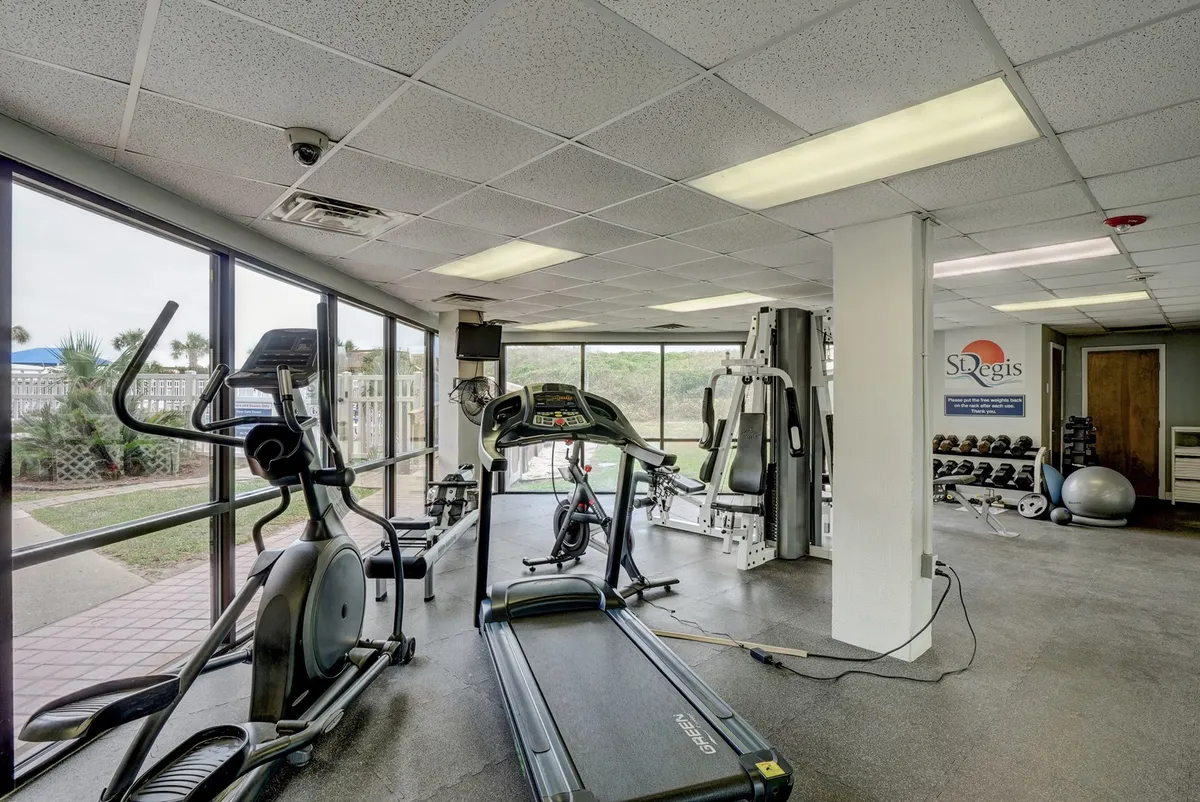 A modern fitness center filled with treadmills, ellipticals, and weight machines is bathed in natural light from large windows overlooking the outdoors. The layout encourages both individual workouts and community fitness activities.
