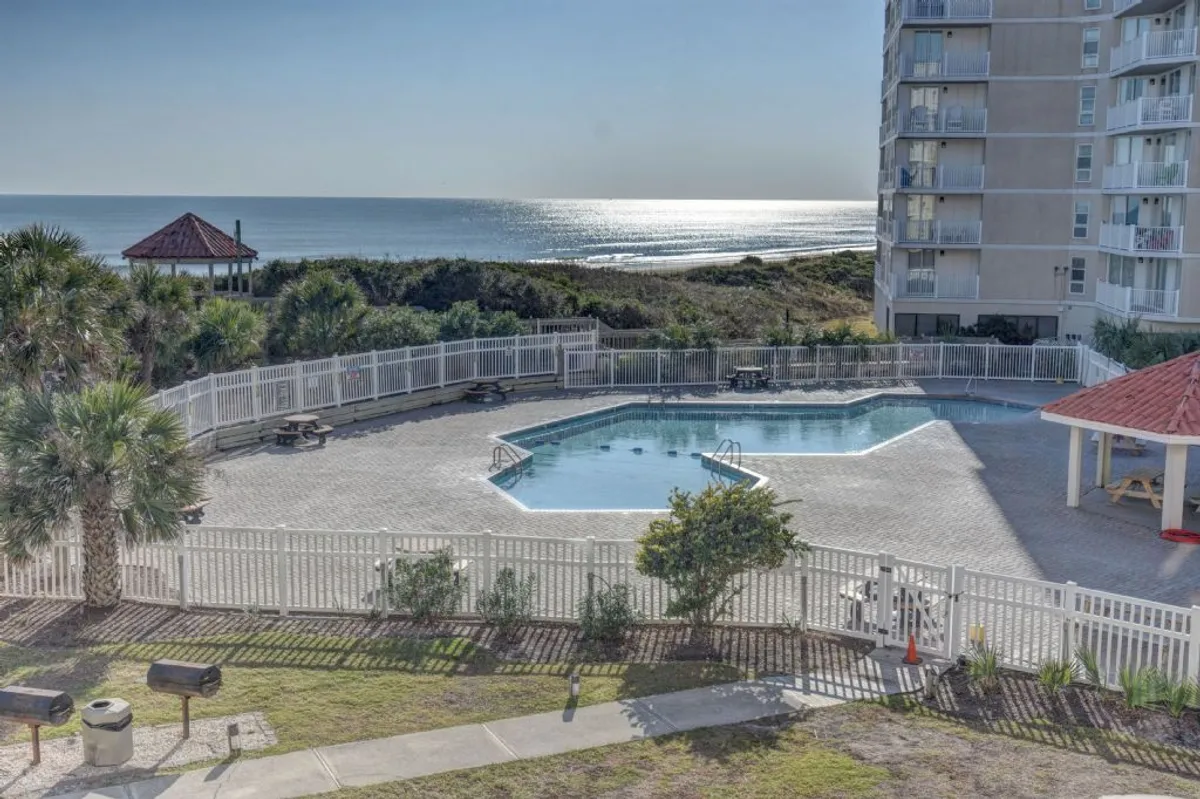 This exterior image showcases a sparkling swimming pool with a charming gazebo, surrounded by landscaped gardens and boasting an inviting view of the ocean in the background. The area is clearly set up for both swimming and outdoor recreation, ideal for families seeking fun and relaxation on-site.