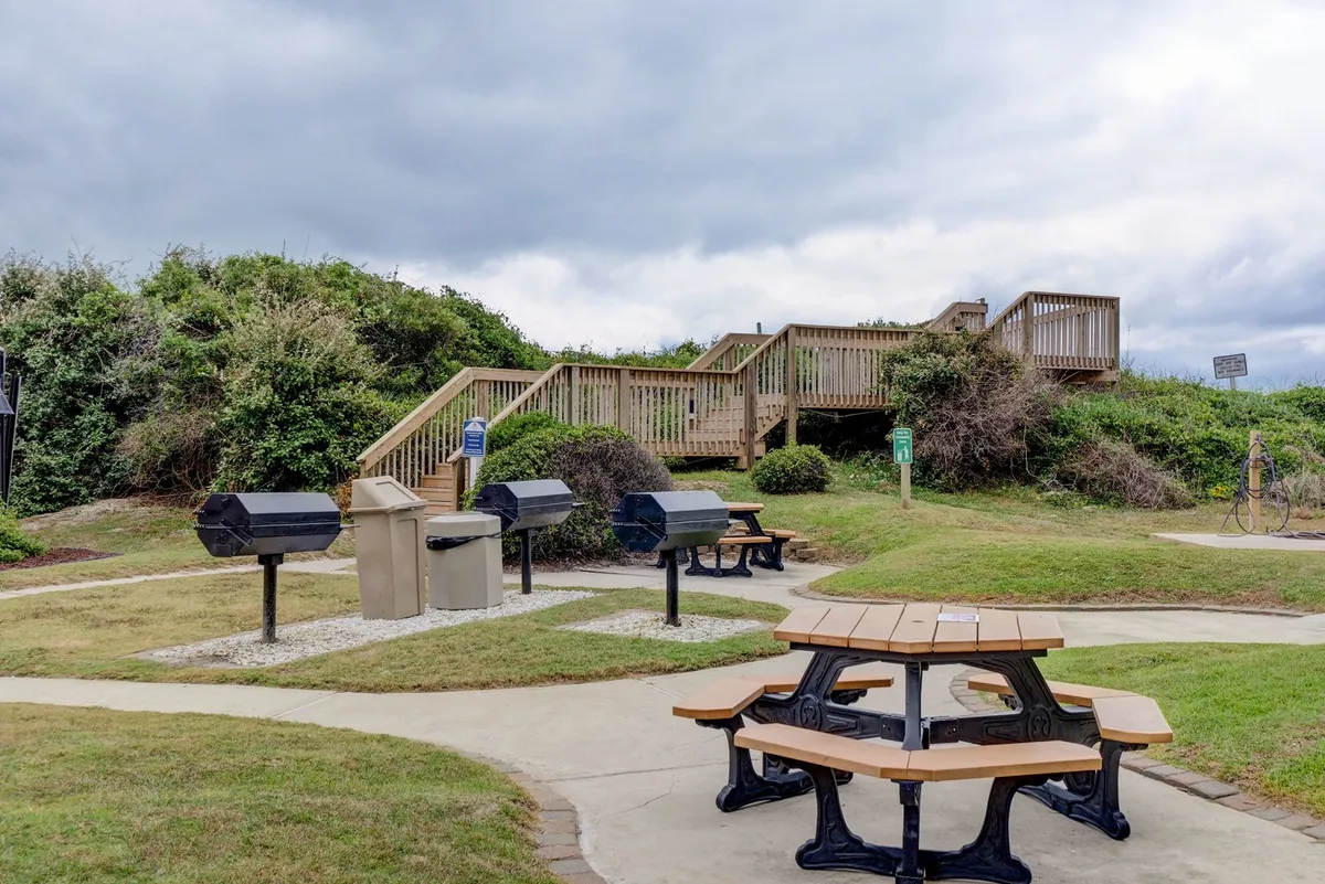An outdoor grilling area with picnic tables and grill stations, set under open skies and adjacent to a staircase, offers a relaxed spot for communal meals and barbecues. The scene is primed for gatherings and shared celebrations.