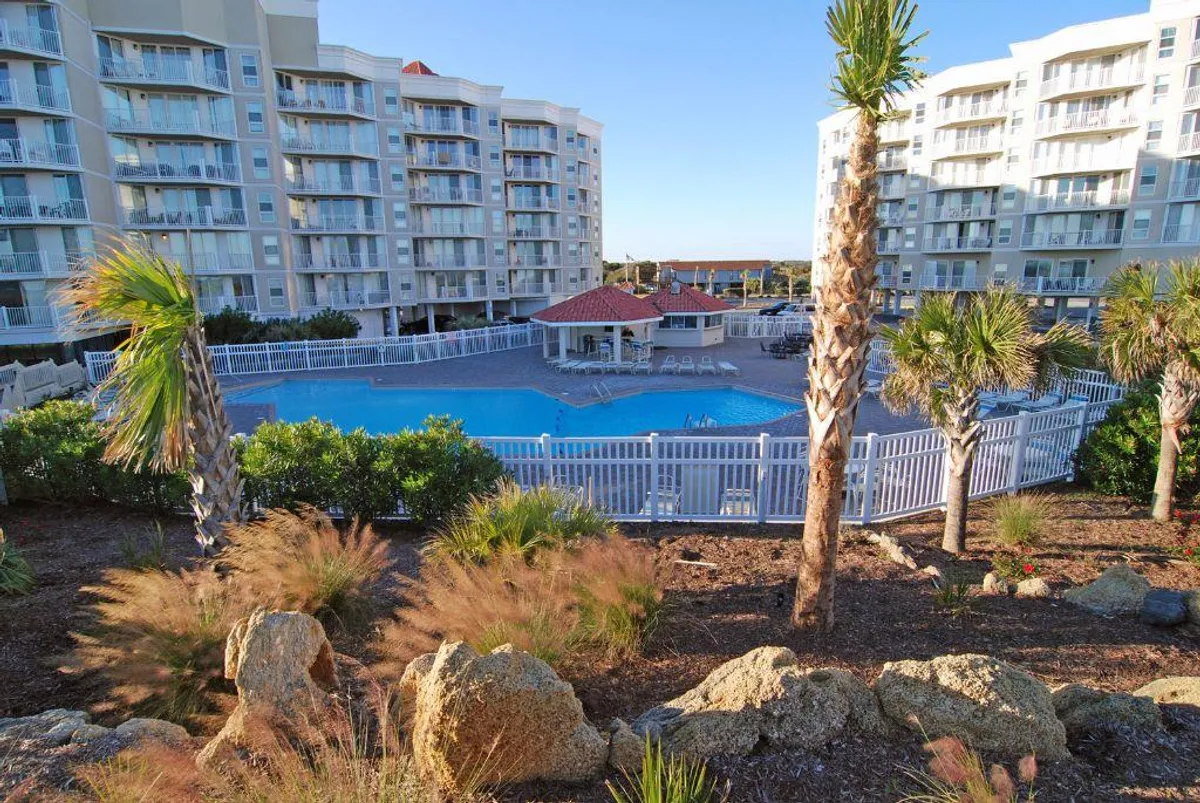 A spacious outdoor swimming pool surrounded by palm trees and landscaped gardens, with the backdrop of resort buildings. Lounge chairs line the pool, inviting guests to relax or socialize. The area is bathed in natural light, evoking a cheerful and leisurely ambiance perfect for gatherings.
