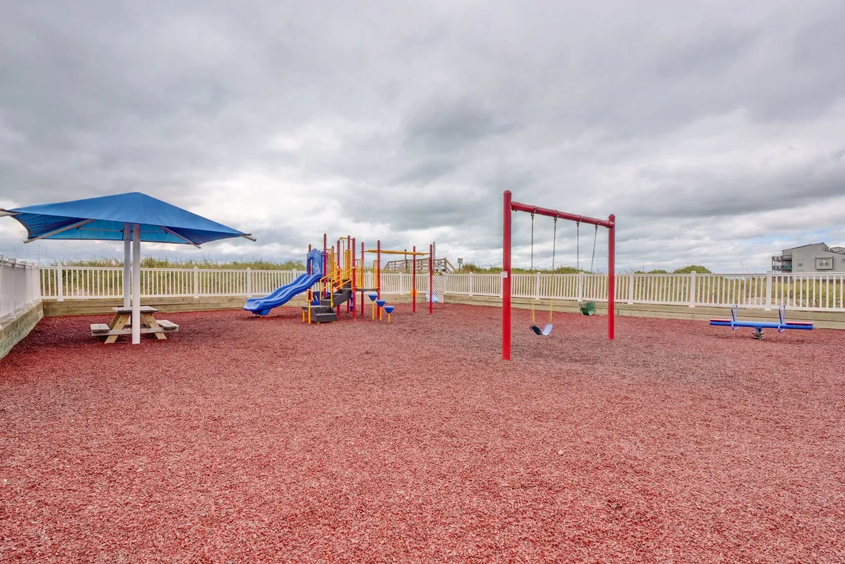 A vibrant outdoor playground with colorful equipment and swings sits under a canopy, adjacent to picnic areas. Children are actively playing, and families are gathered nearby, creating a cheerful, community-focused atmosphere.