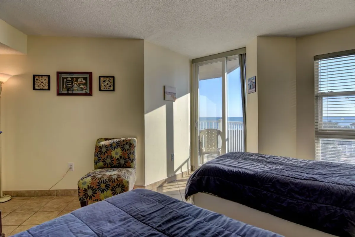 A cozy bedroom featuring two beds, an armchair by the window, and a balcony view that hints at the nearby ocean. The space is inviting, ideal for families or friends sharing the retreat.