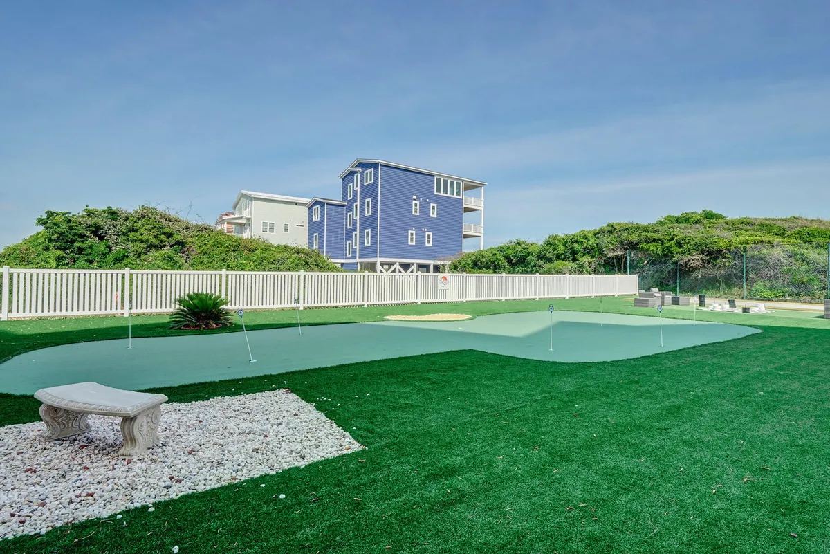 A well-maintained putting green set against a landscaped area with a blue house in the distance. The green is inviting and ready for friendly competition or relaxed practice in the sunshine.
