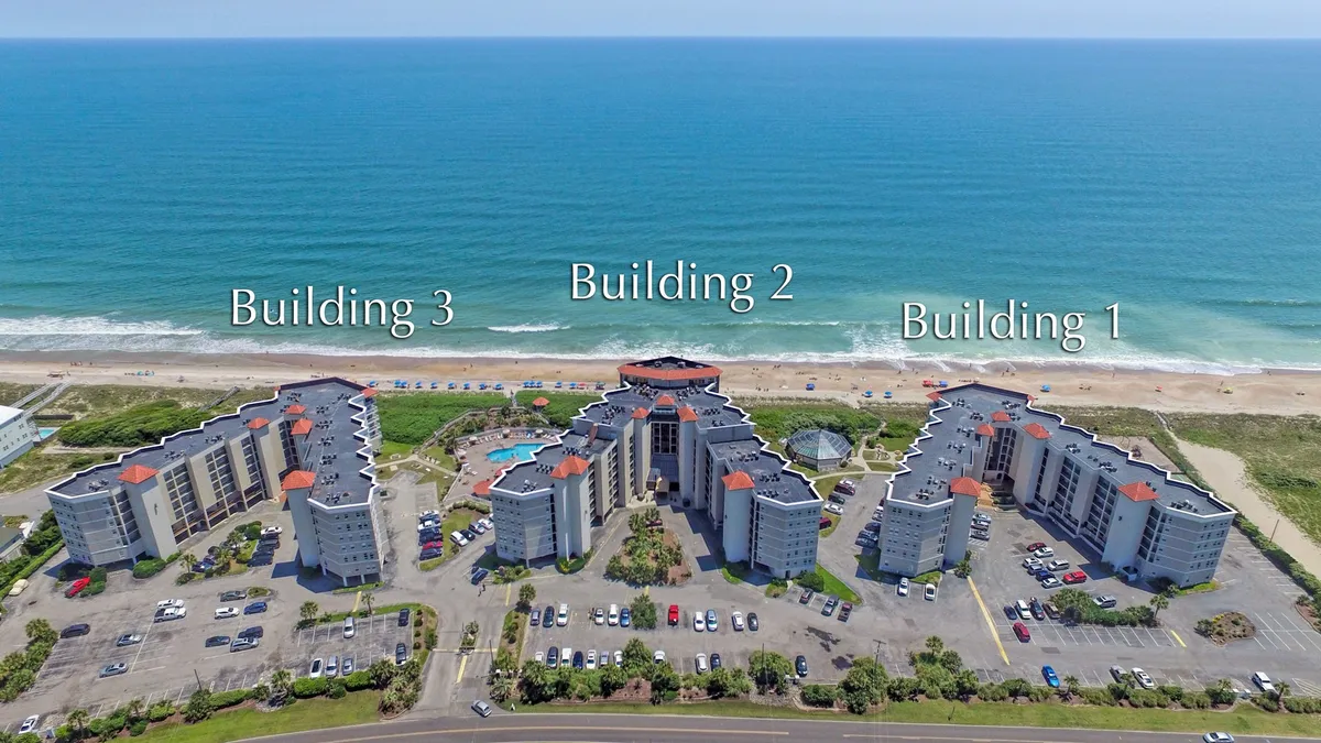 An elevated exterior view showcases three modern buildings flanking a large swimming pool, with the vast ocean stretching into the distance. The scene features spaces for swimming, sunbathing, and walking along the beach, all set against a serene coastal backdrop ideal for wellness and relaxation.