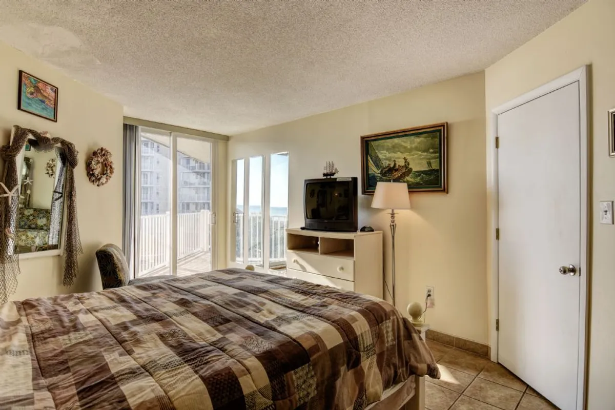 A tranquil bedroom with a patterned quilt on the bed, large sliding glass doors that open to reveal ocean views, a TV unit, and a decorative mirror. The space feels airy and private, perfect for unwinding after a day at the beach.