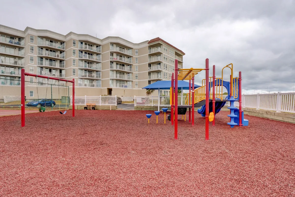 A vibrant playground fitted with swings and modern equipment sits atop a soft red rubber surface. Children are seen playing and families gather nearby, creating a lively, community-oriented setting.