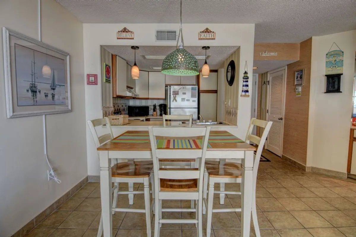 A cozy dining area with a large table, colorful table runner, and an attractive hanging light fixture. The space is full of natural light, offering a welcoming spot for family meals, celebrations, or casual gatherings. Wall decor adds to the inviting atmosphere.
