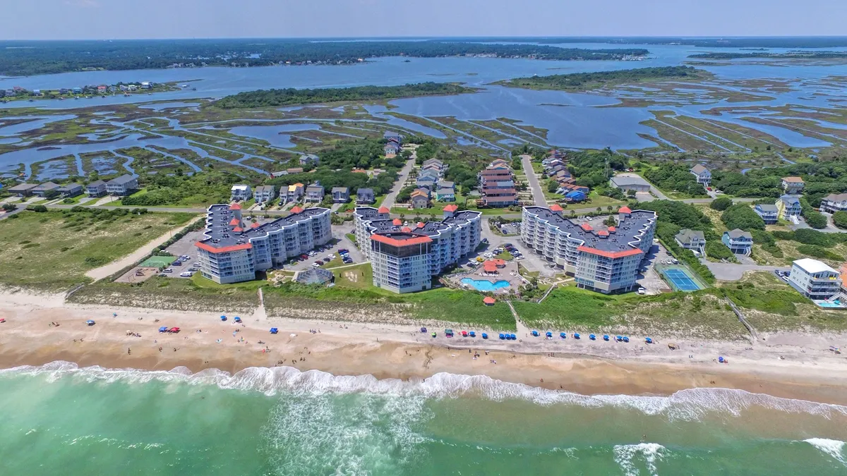This exterior shot captures several beachfront properties with the shimmering swimming pool in the foreground, gentle ocean waves beyond, and colorful beach umbrellas dotting the landscape. The scene exudes relaxation, with people sunbathing, swimming, and enjoying the scenic coastline, perfectly illustrating the romantic and leisurely atmosphere of North Topsail Beach.